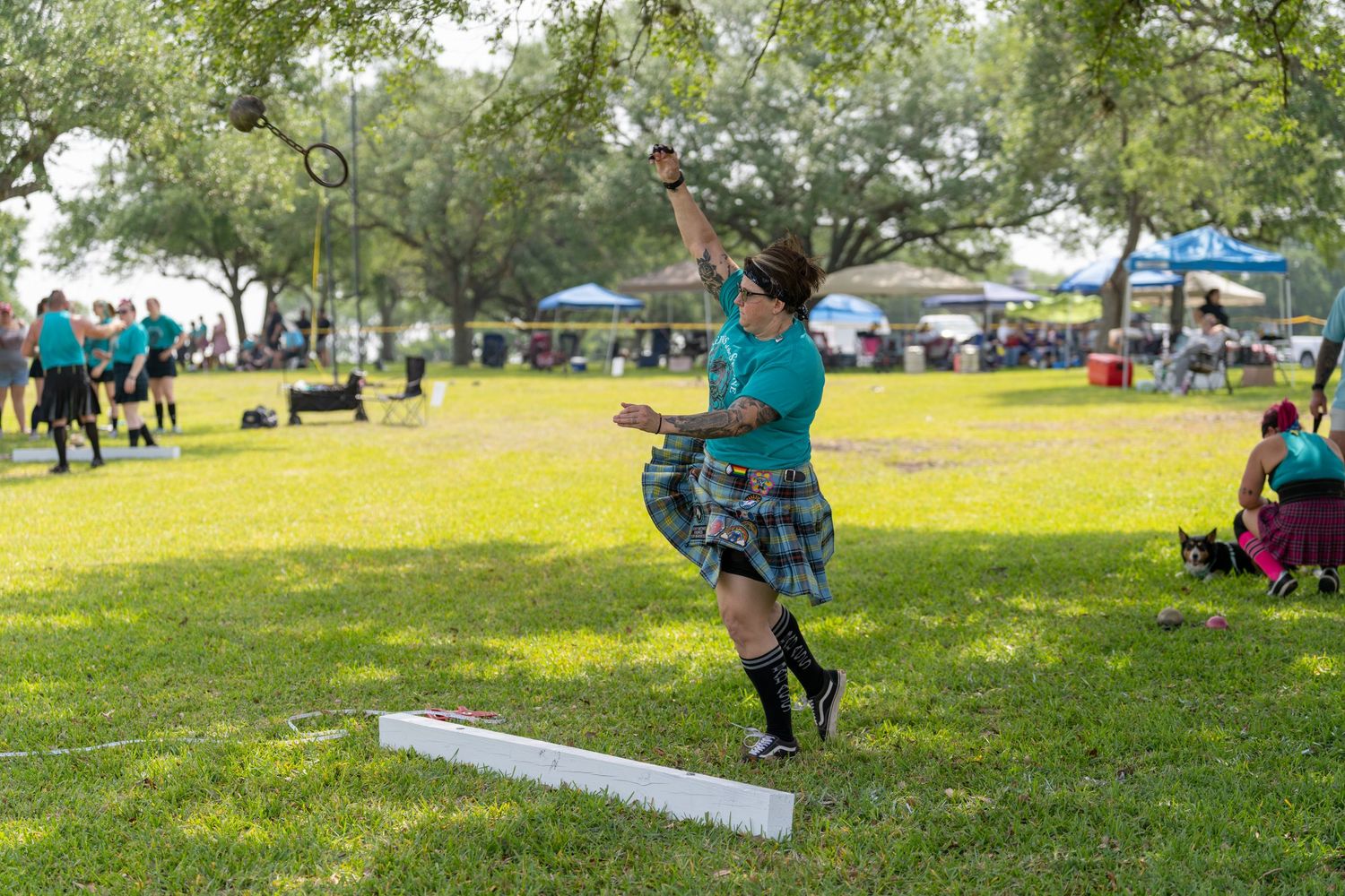 Athlete in tartan kilt performs Highland Games hammer throw on green grass at outdoor Scottish festival.