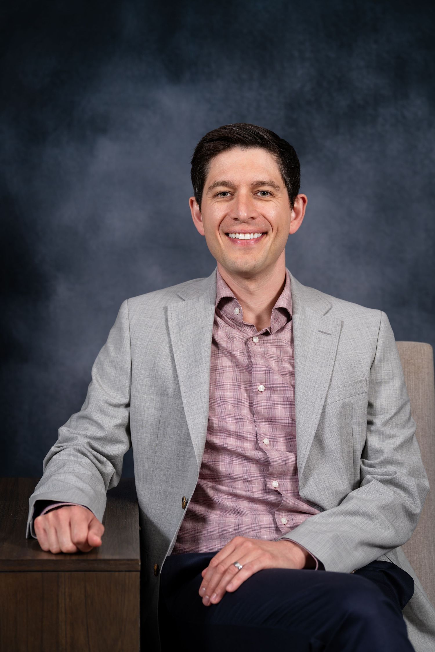 Smiling man in gray blazer and pink plaid shirt seated in a chair against a dark studio background.