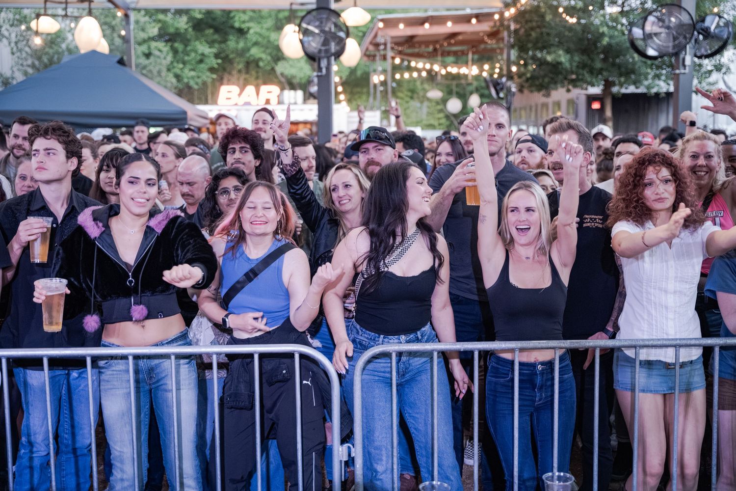Crowd of concert fans behind metal barricades holding beers at an outdoor bar event at night.