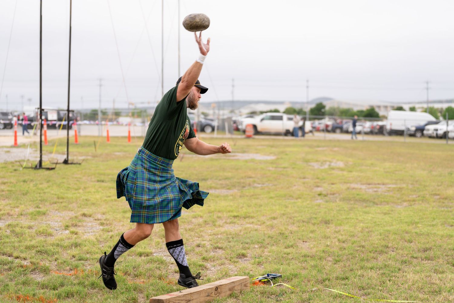 Man in tartan kilt throwing a stone at a Scottish Highland Games competition outdoors.