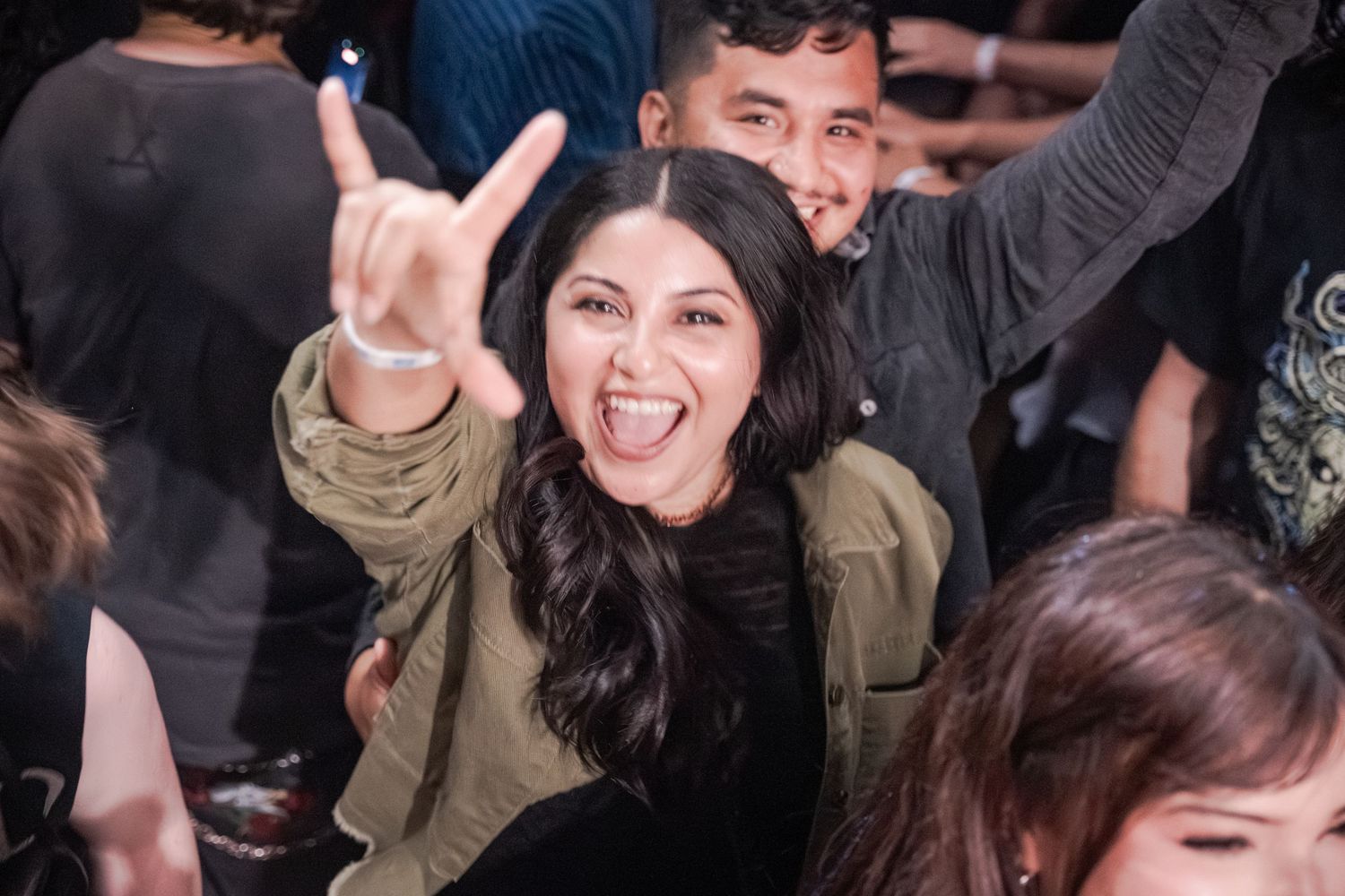 Excited woman flashing rock on hand sign at a crowded concert, wearing olive jacket with long dark hair.