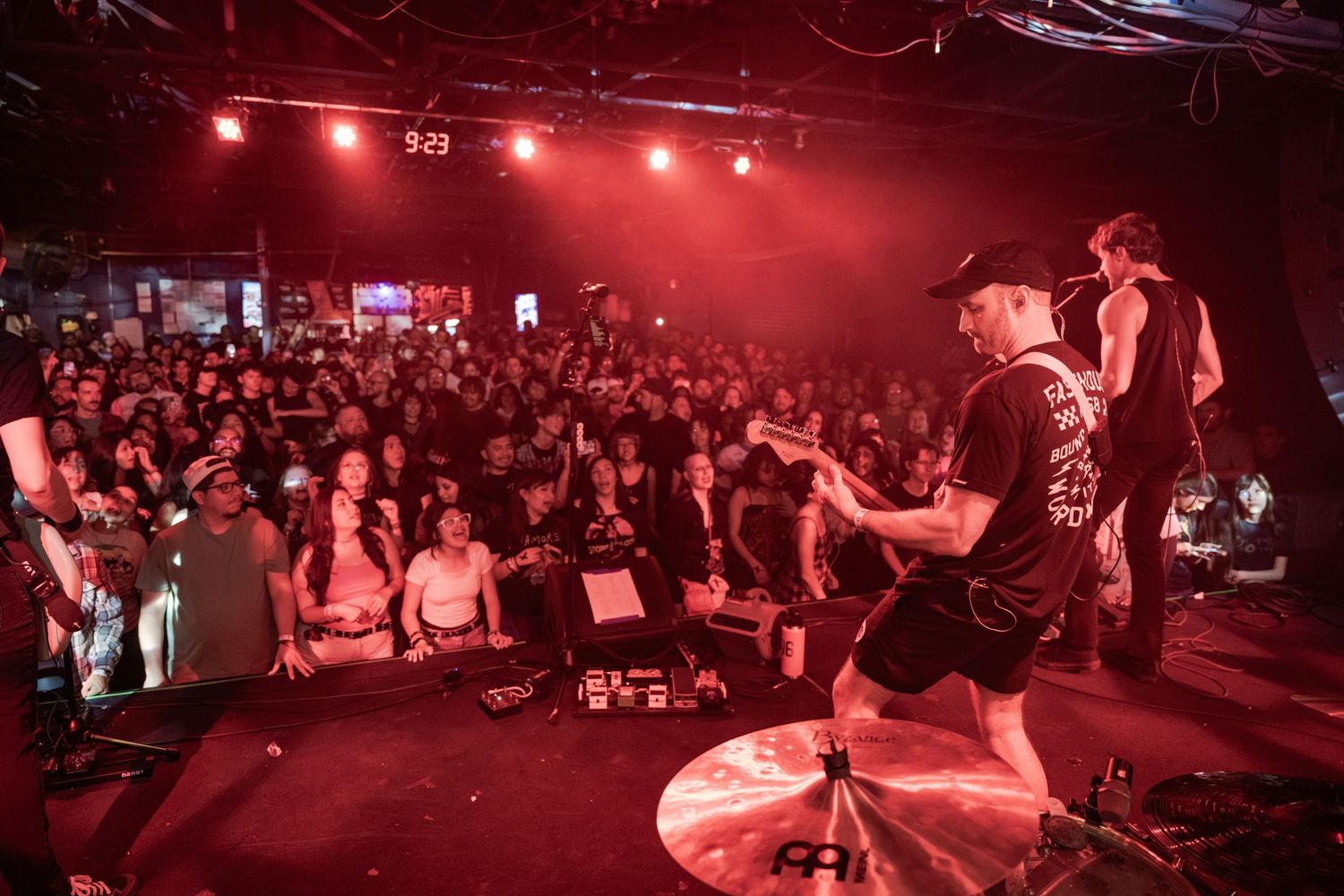 Band performing on stage under red lights before a large crowd at an indoor rock concert venue.