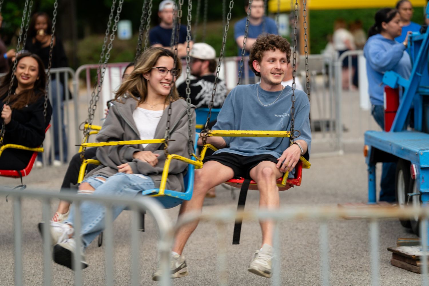 Two young adults laughing and enjoying a swing ride at an outdoor amusement park or fair on a cloudy day.