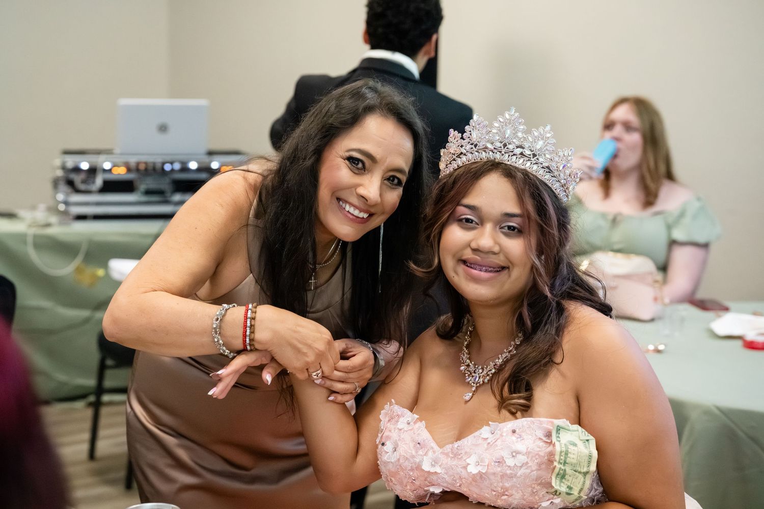 Two women pose together at a quinceañera celebration, one wearing a sparkling tiara and pink gown.
