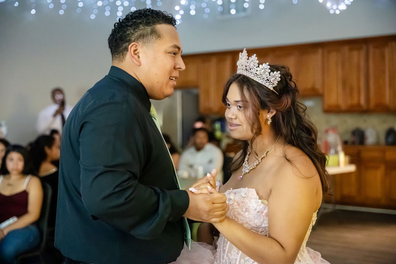 A young woman in a tiara and pink gown shares a dance with a man in a dark suit at a quinceañera.