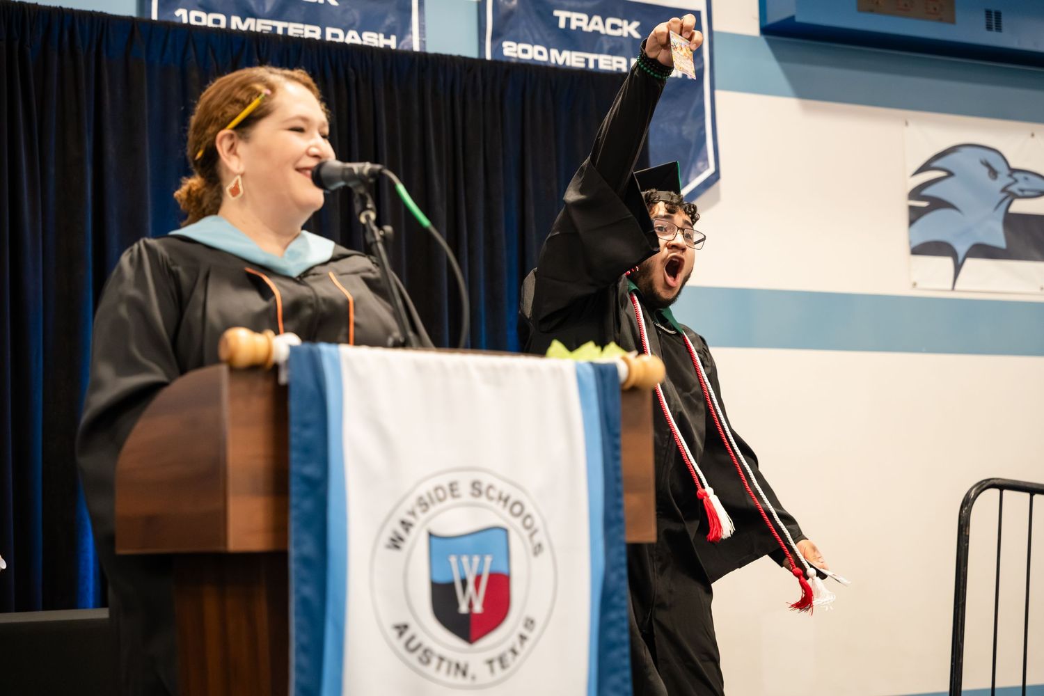 Graduate celebrates joyfully at Wayside School Austin Texas graduation ceremony beside speaker at podium.