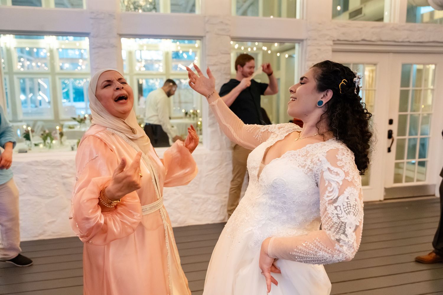 Bride in white lace gown dancing joyfully with a guest in peach hijab at an elegant wedding reception.