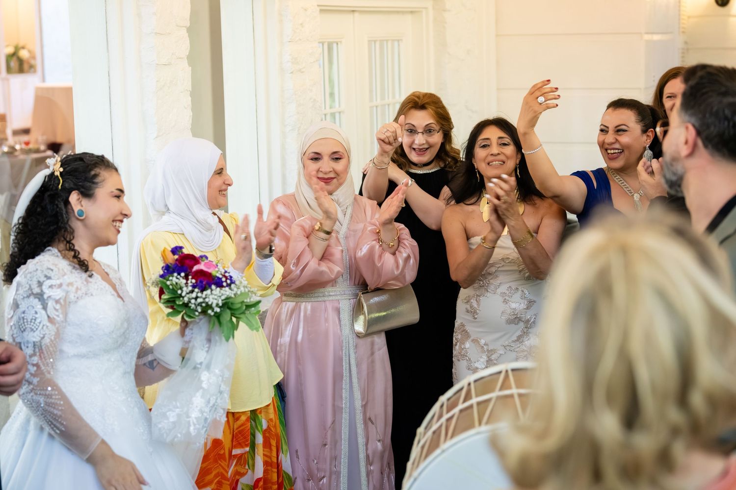 Bride in white gown holds colorful bouquet surrounded by celebrating guests at elegant wedding reception.