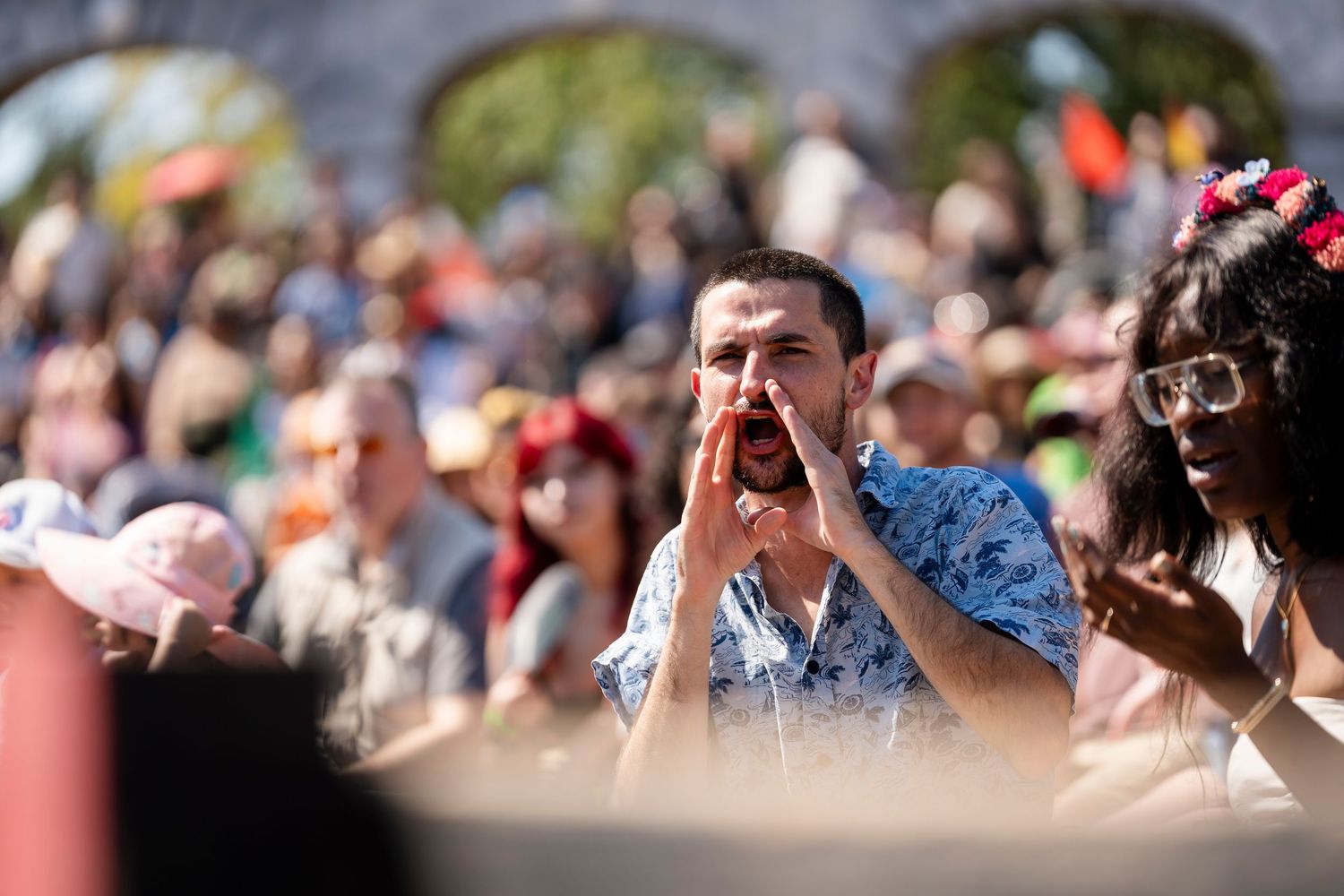 A large crowd of spectators watches an outdoor event with people visible in the background on a sunny day.