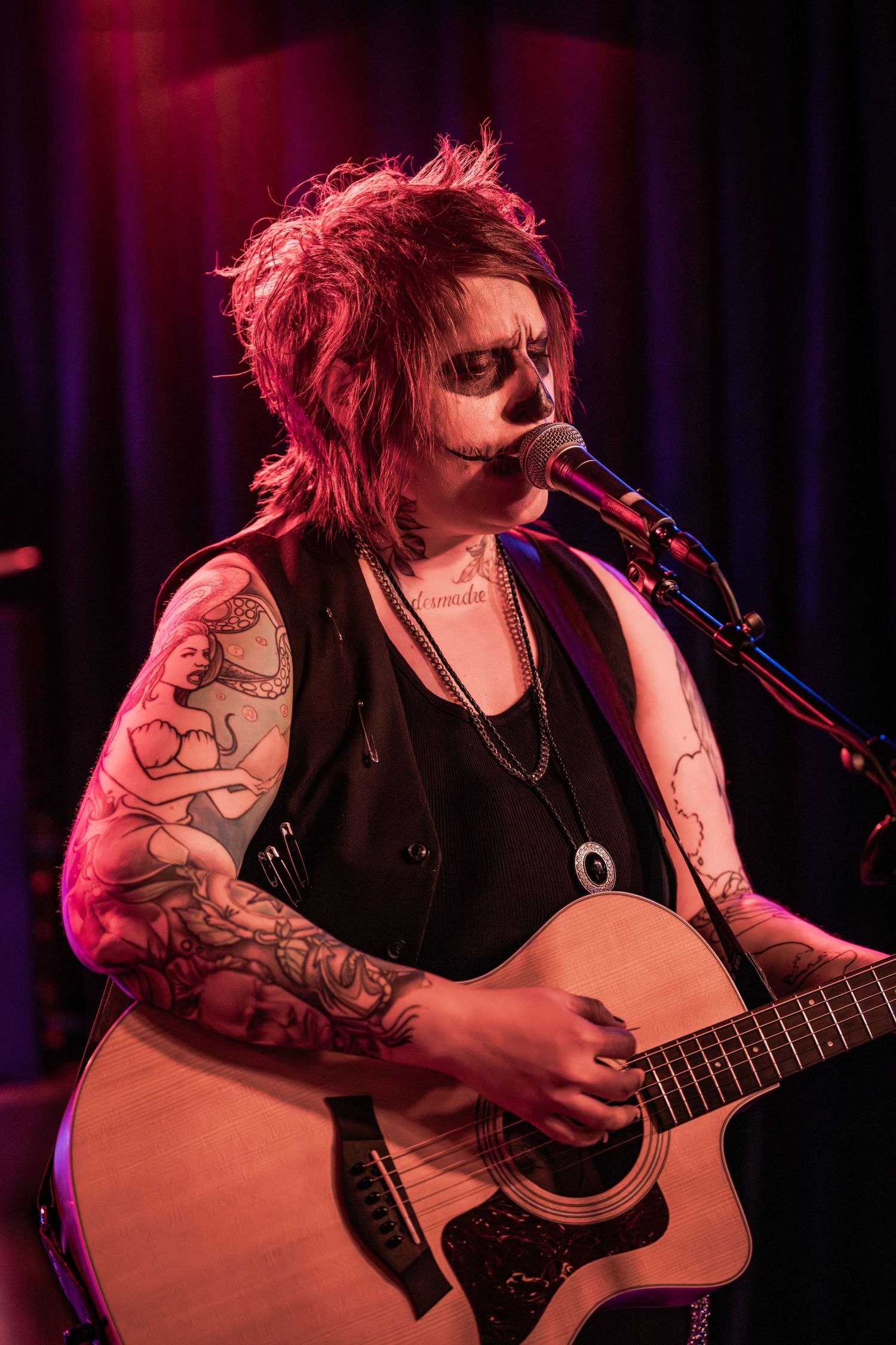 A musician performs on acoustic guitar under dramatic red stage lighting during an intimate concert performance.