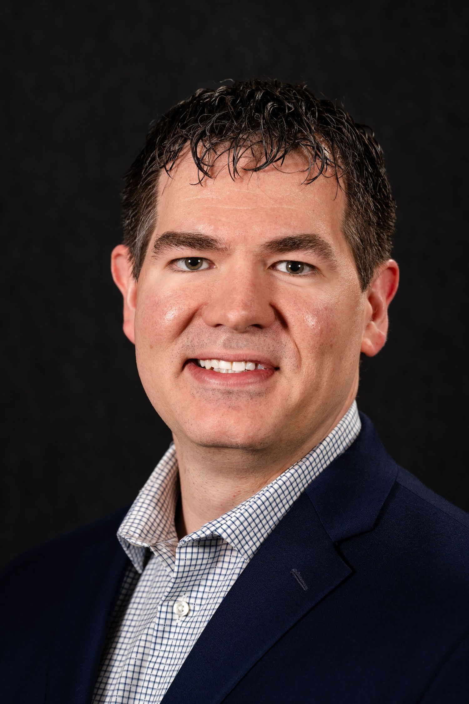 Professional headshot of person in navy blazer and checkered dress shirt against dark background.