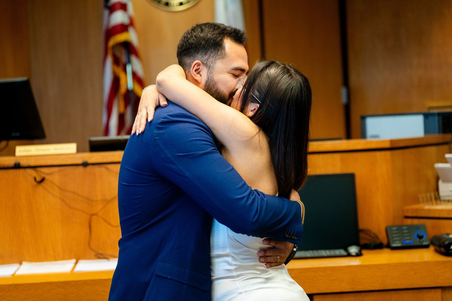 A couple shares a romantic kiss in a courthouse setting with warm lighting and wooden decor.