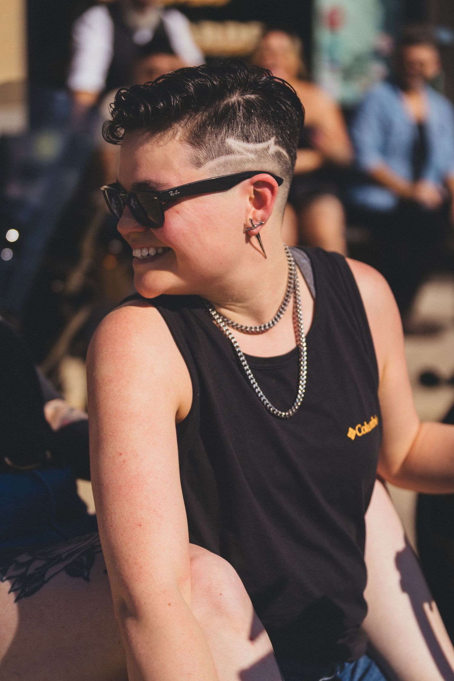 A person in a black tank top and sunglasses smiles while wearing silver chain necklaces and an undercut hairstyle.