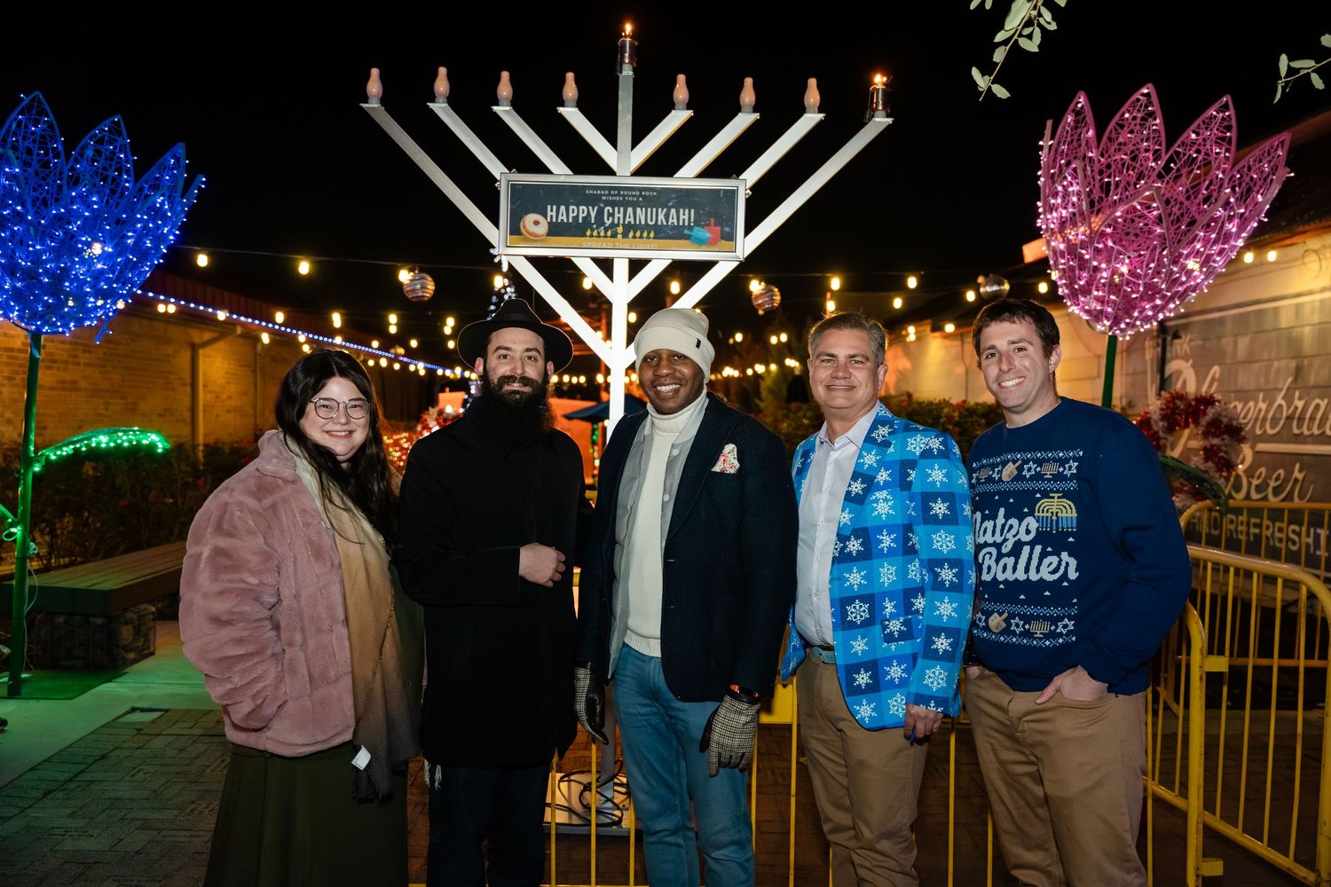 People gather around outdoor holiday displays featuring illuminated Hanukkah menorahs and colorful lights at night.