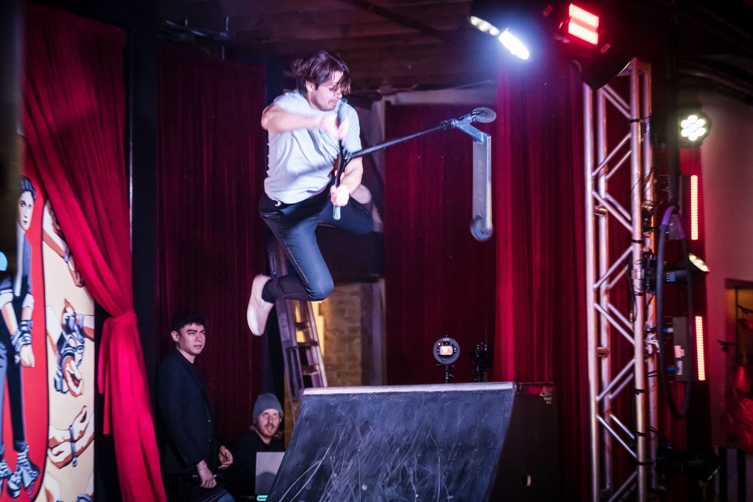 A performer jumps high off a platform on stage with red curtains and stage lighting in the background.