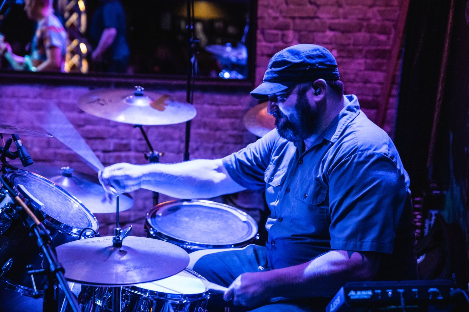 A drummer performs on stage with purple and blue lighting illuminating the drum kit and cymbals during a live performance.