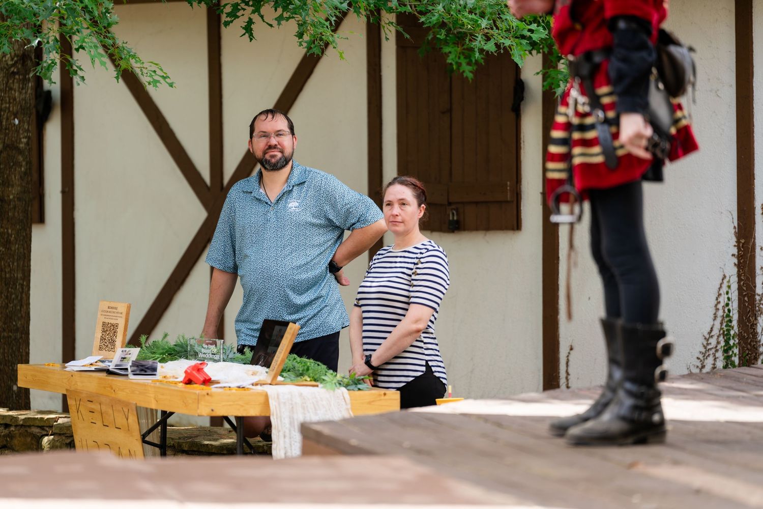 Two people stand behind a vegetable market stall outdoors near a rustic building, with a third person visible in the background.
