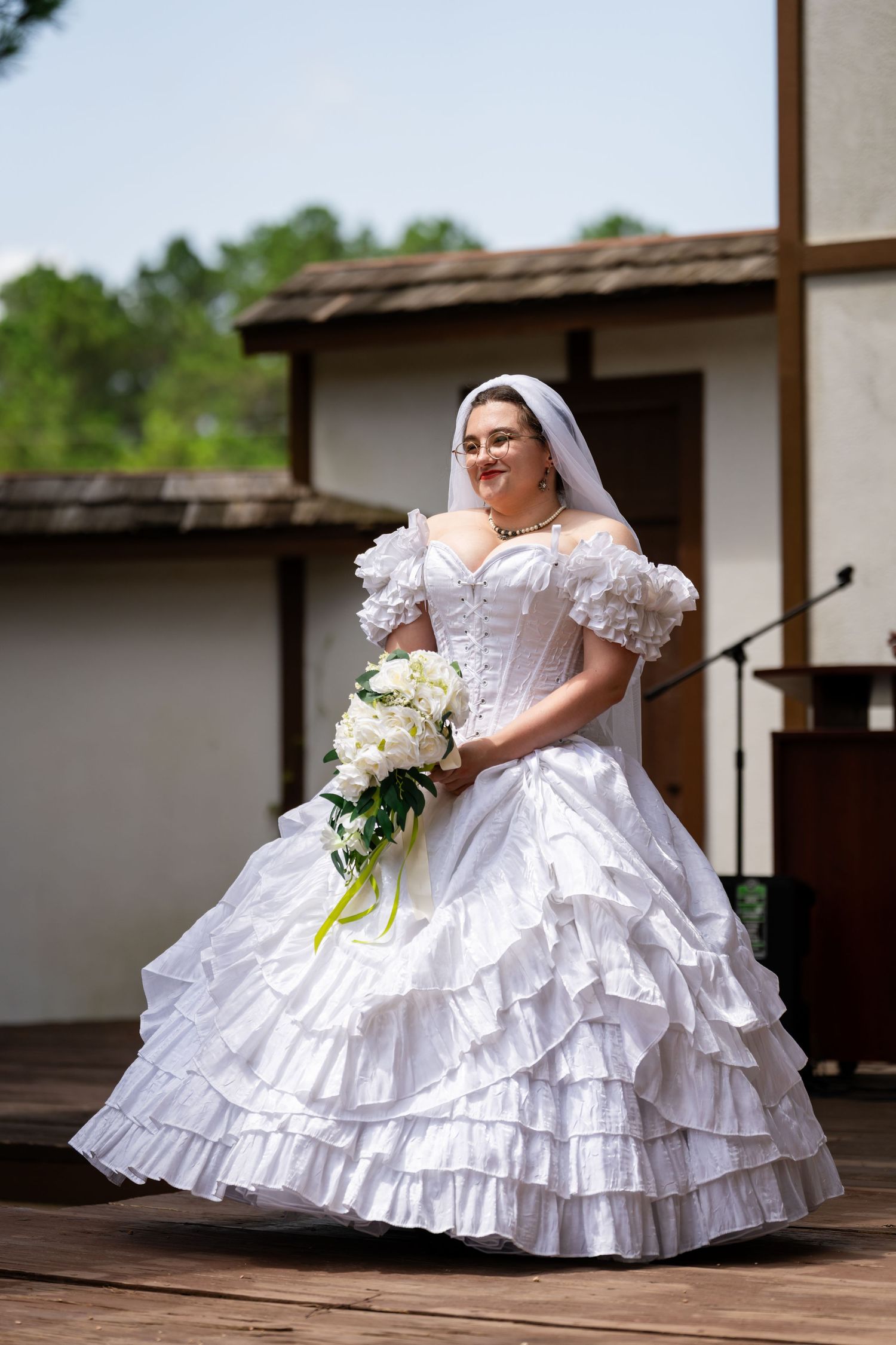 Woman in white Victorian bridal gown with ruffled skirt, veil, and bouquet of white flowers on outdoor stage.
