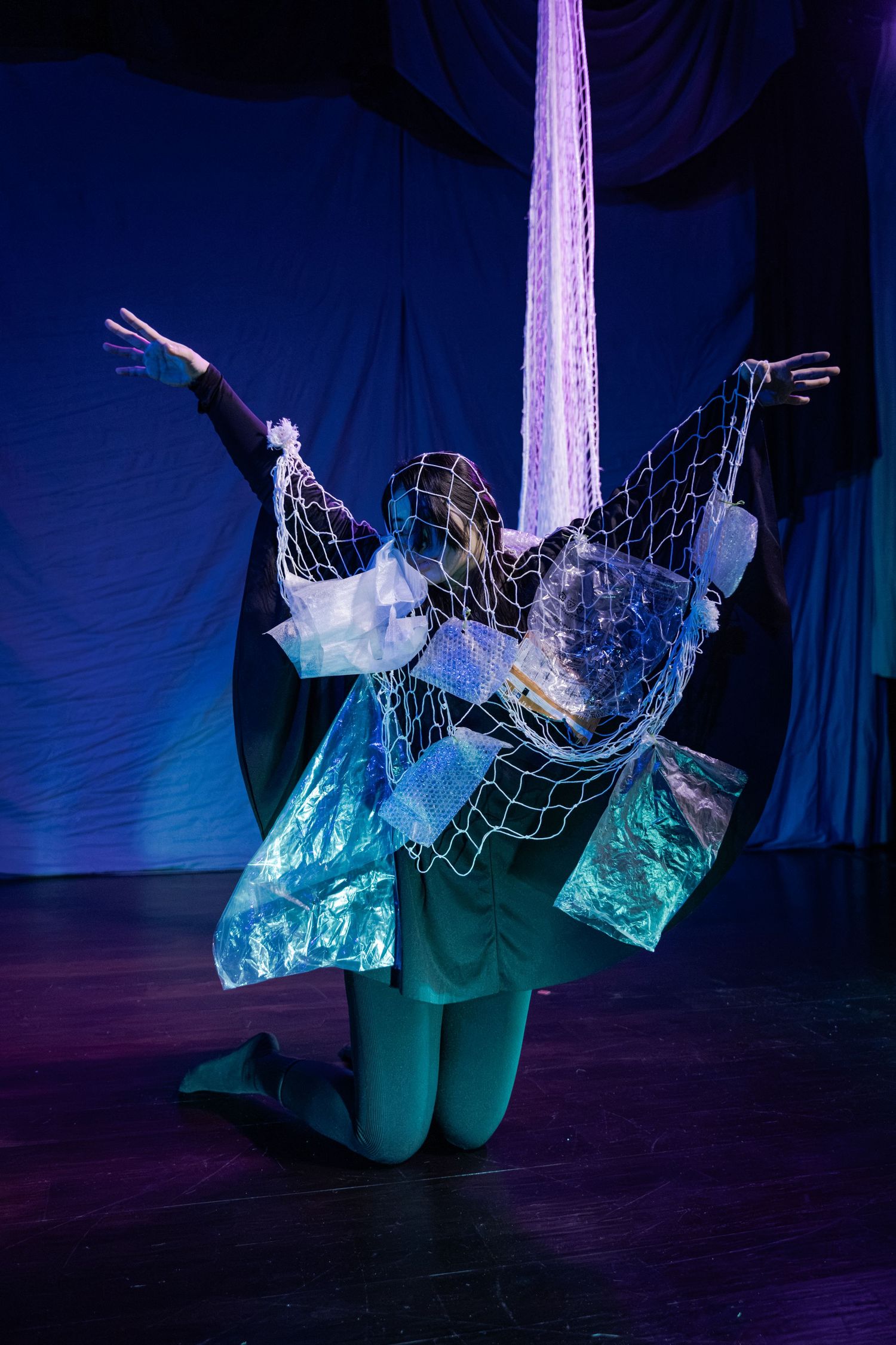 Performer in aquatic costume with netting and blue plastic, kneeling on stage under purple aerial silk.