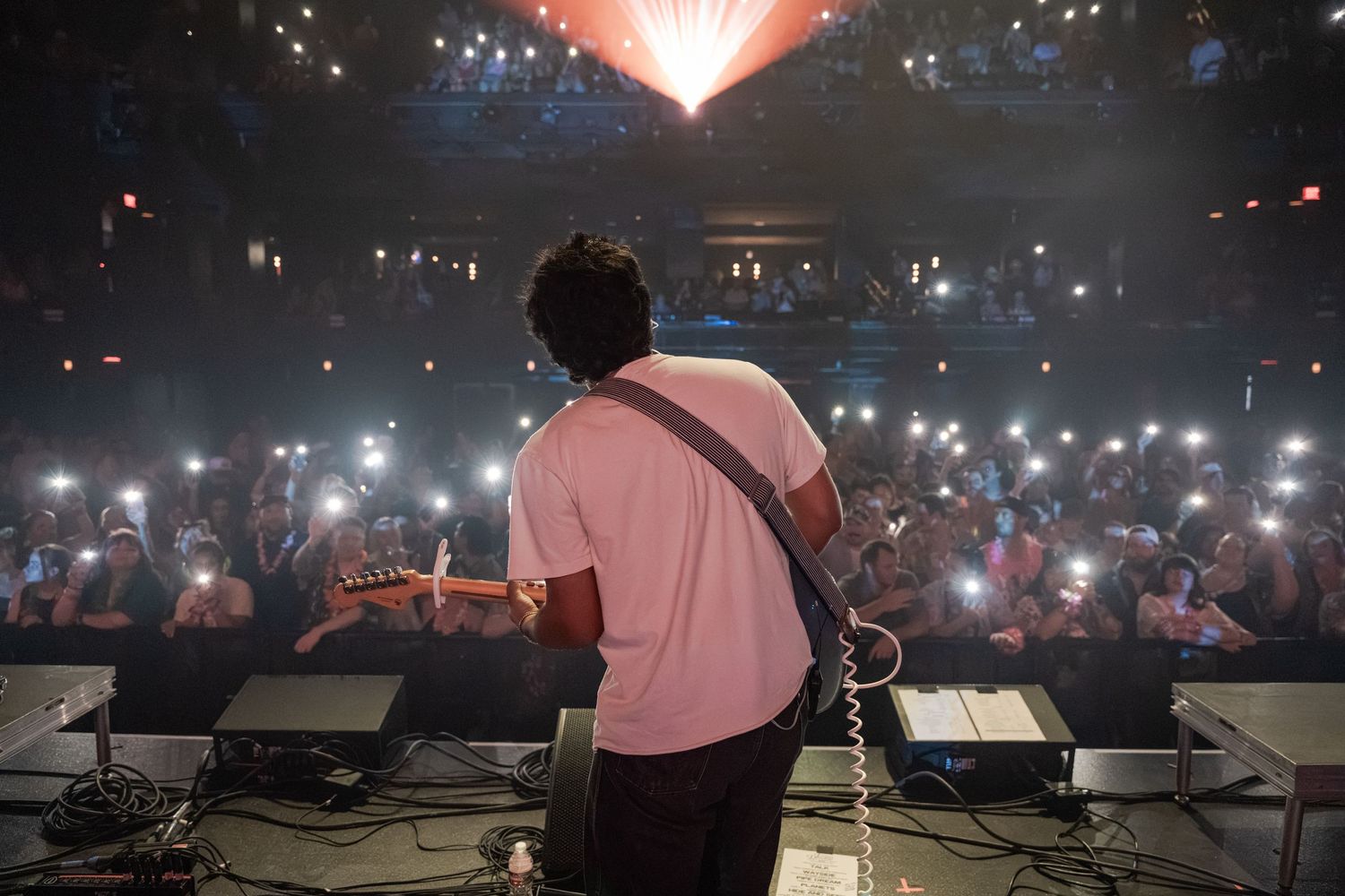 Guitarist performs on stage with back to camera as crowd holds up phone flashlights in a packed concert venue.