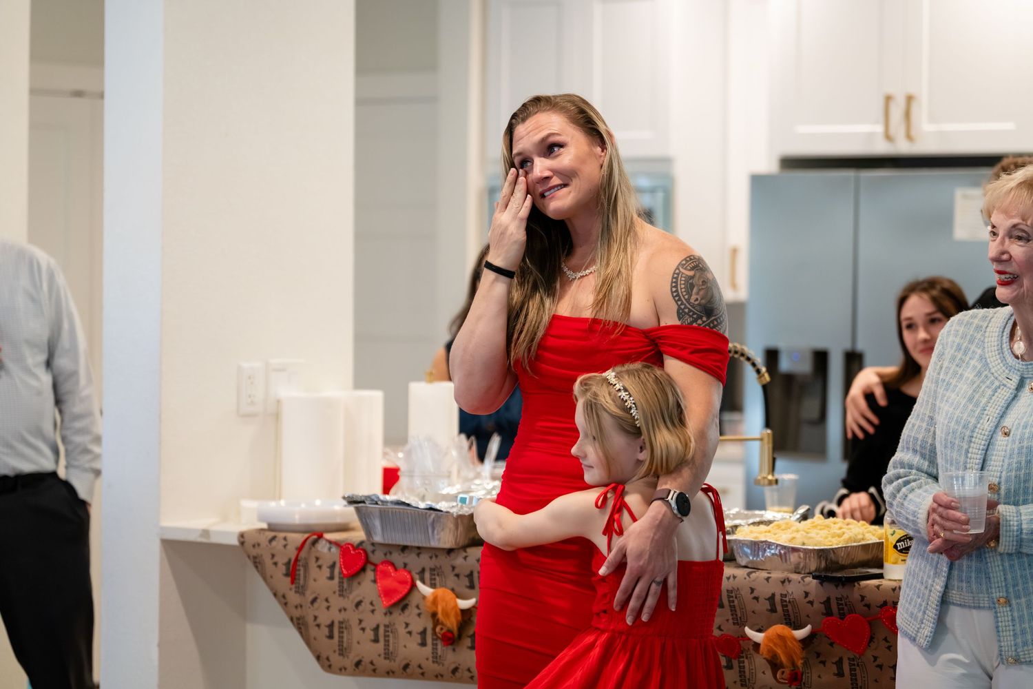 Woman in red dress crying while hugging a young girl at an indoor celebration with guests in background.