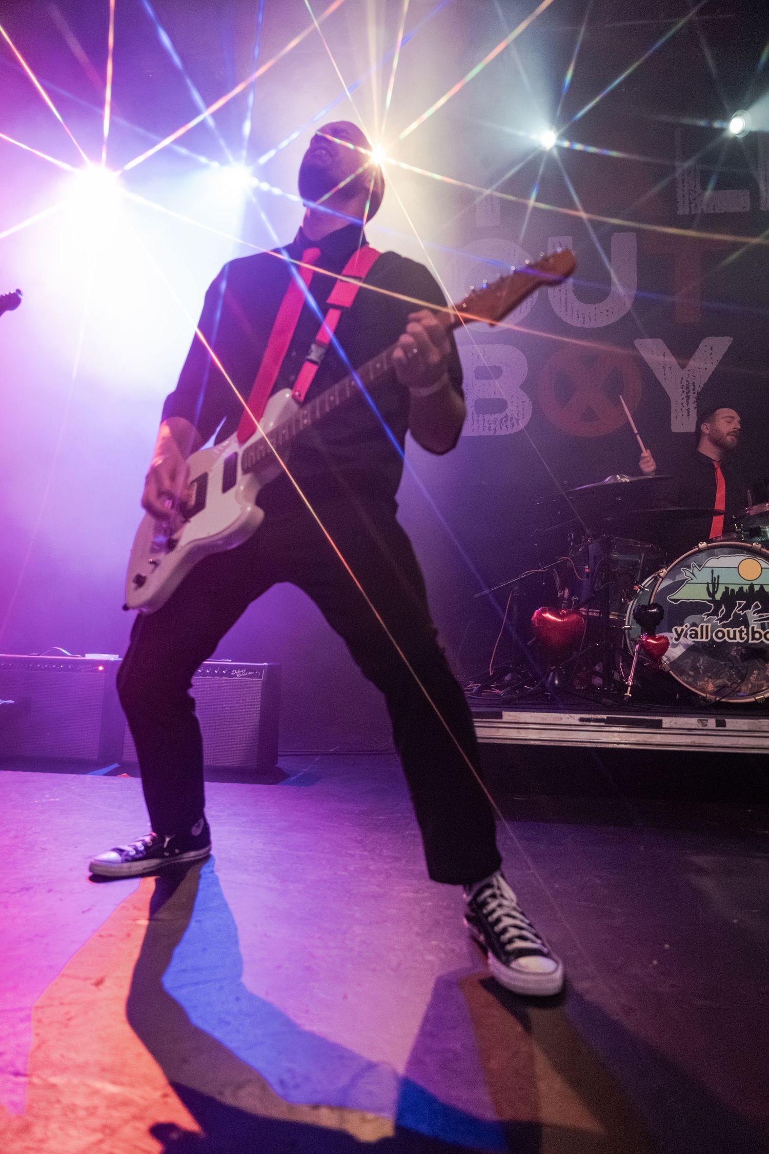 Guitarist performing on stage with dramatic lighting, wearing black outfit and Converse sneakers, playing white electric guitar.