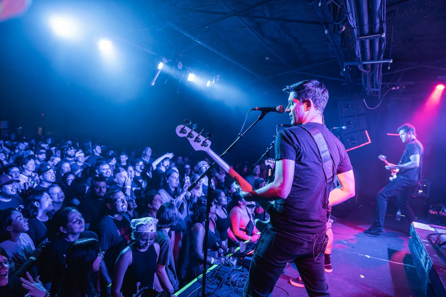 A musician performs on stage with a bass guitar under blue and pink concert lighting while a crowd watches in the foreground.