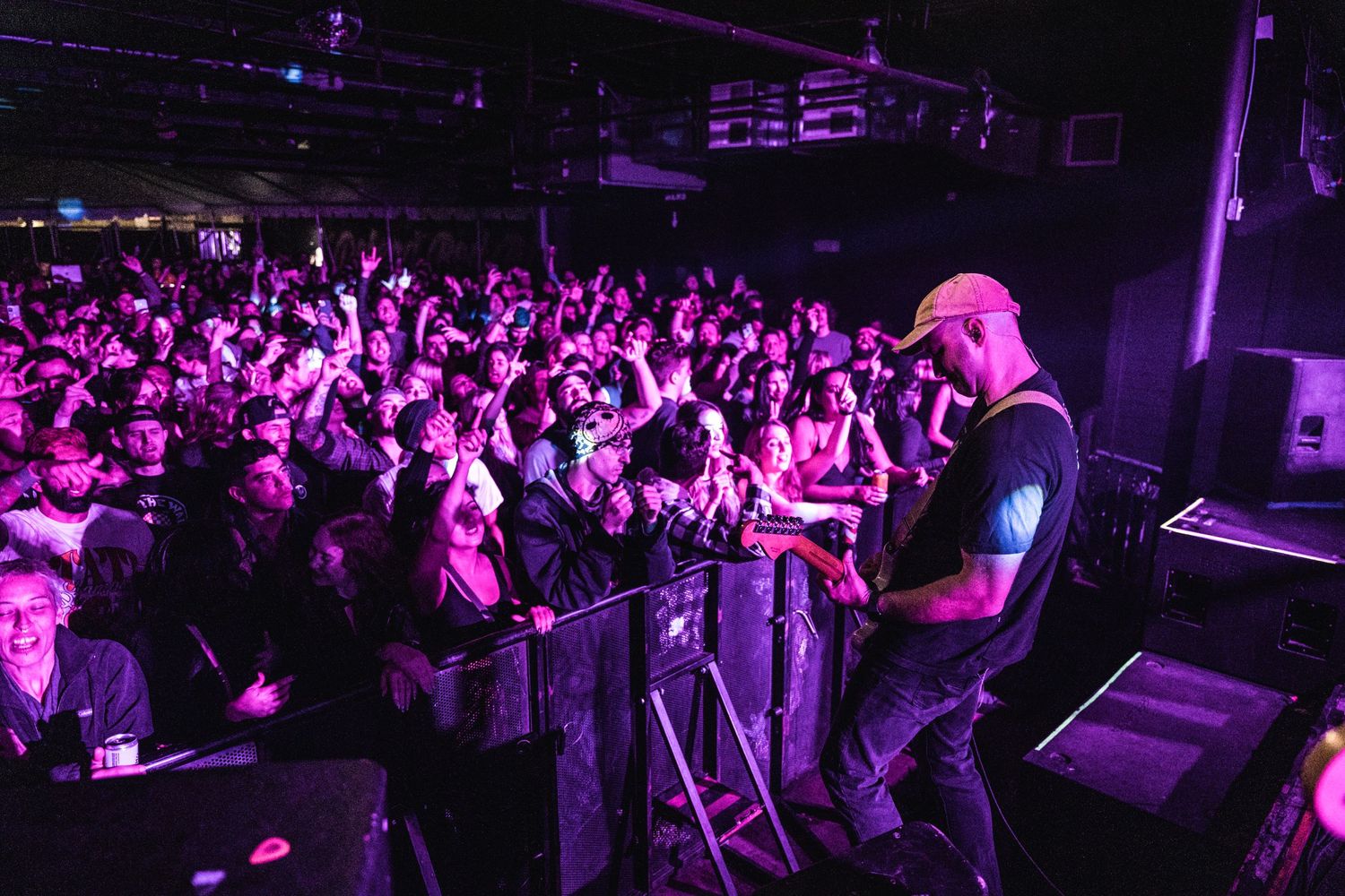 Enthusiastic crowd at a rock concert illuminated by purple lights while performer plays on stage.