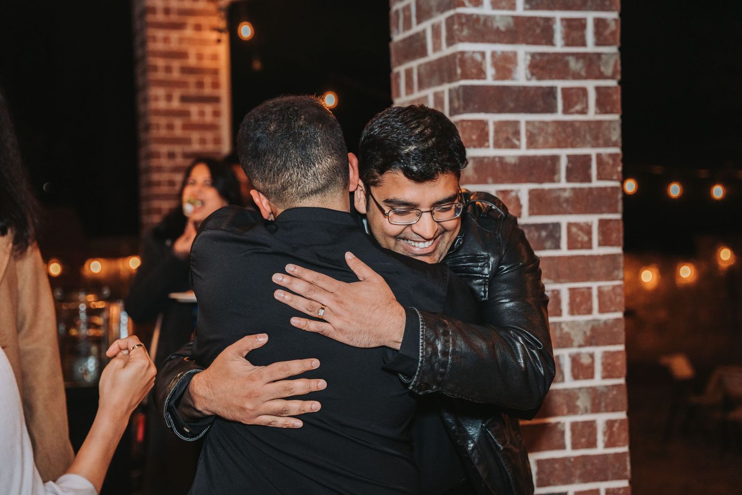 Emotional embrace captured at celebration against brick wall with string lights.