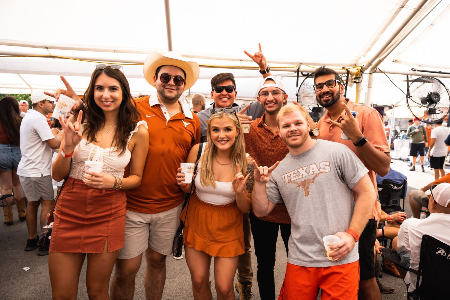 A group of sports fans wearing burnt orange and white gather under a tent at a tailgate party.