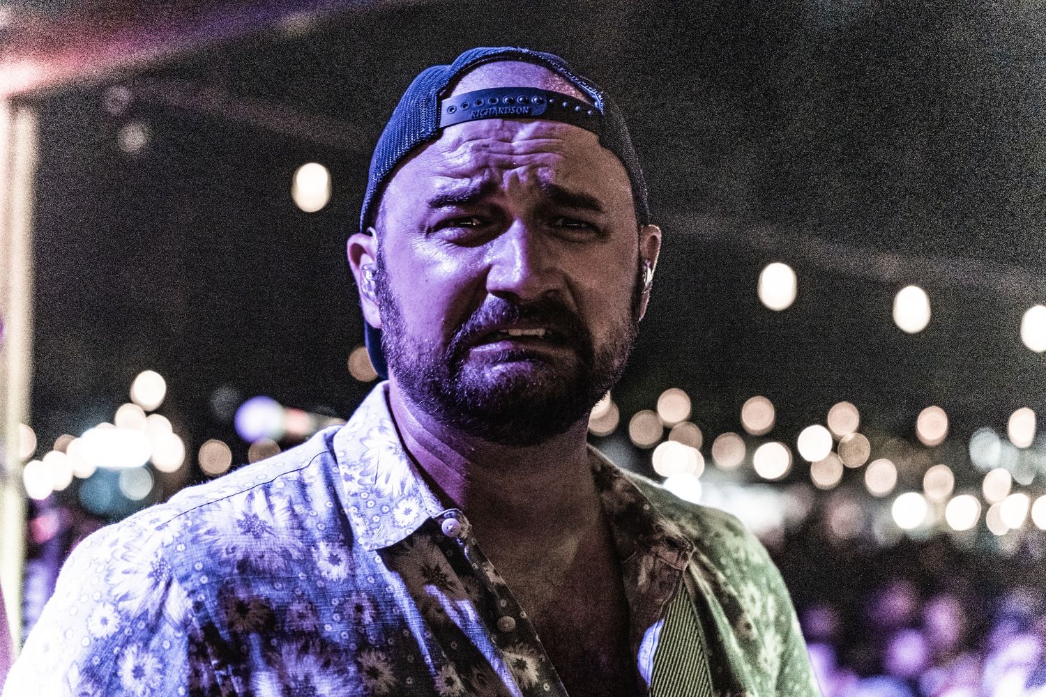 Performer in a patterned shirt and cap singing on stage with stage lights in background.