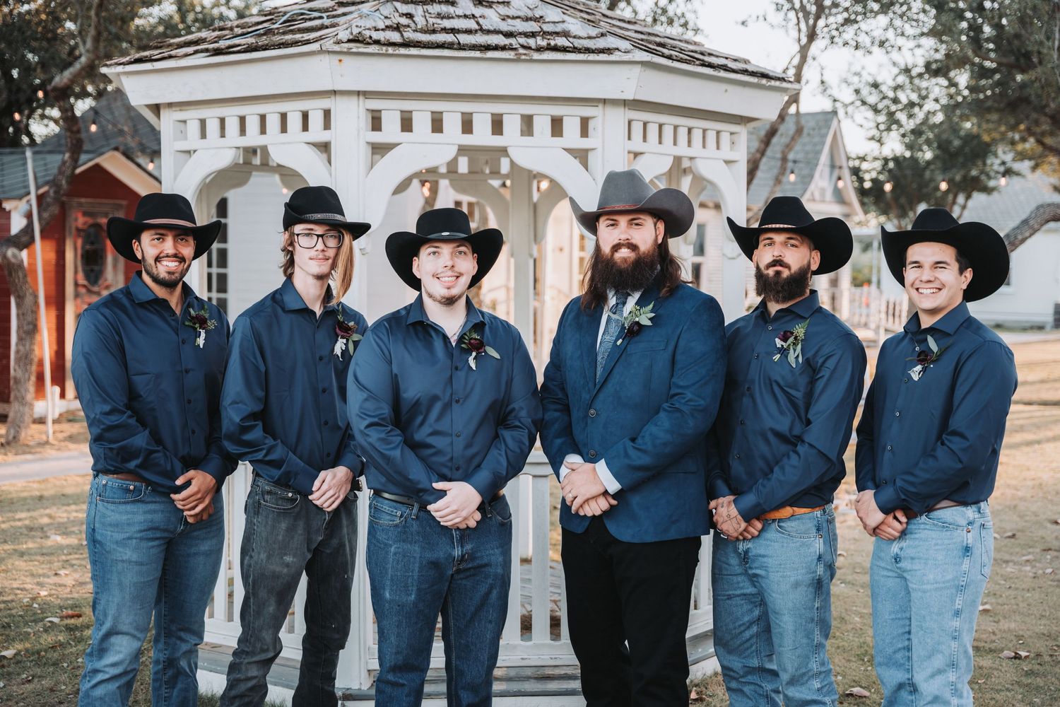 Groomsmen in matching blue shirts and cowboy hats pose in front of a white gazebo.