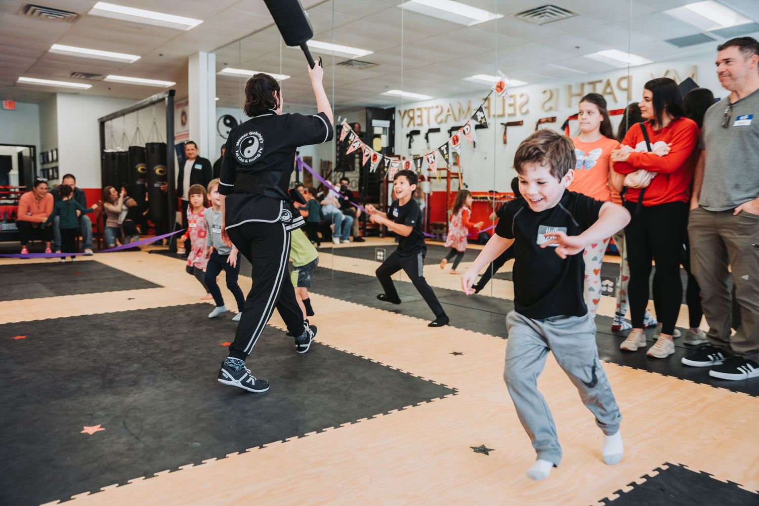 Students practice martial arts moves on floor mats while others observe along the wall.