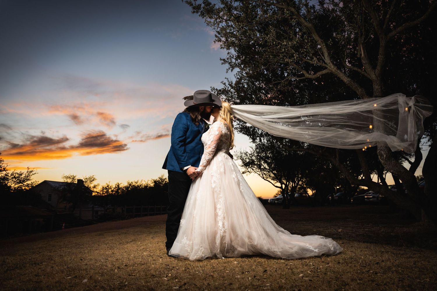 Couple in wedding attire posing against sunset with flowing veil in dramatic lighting.