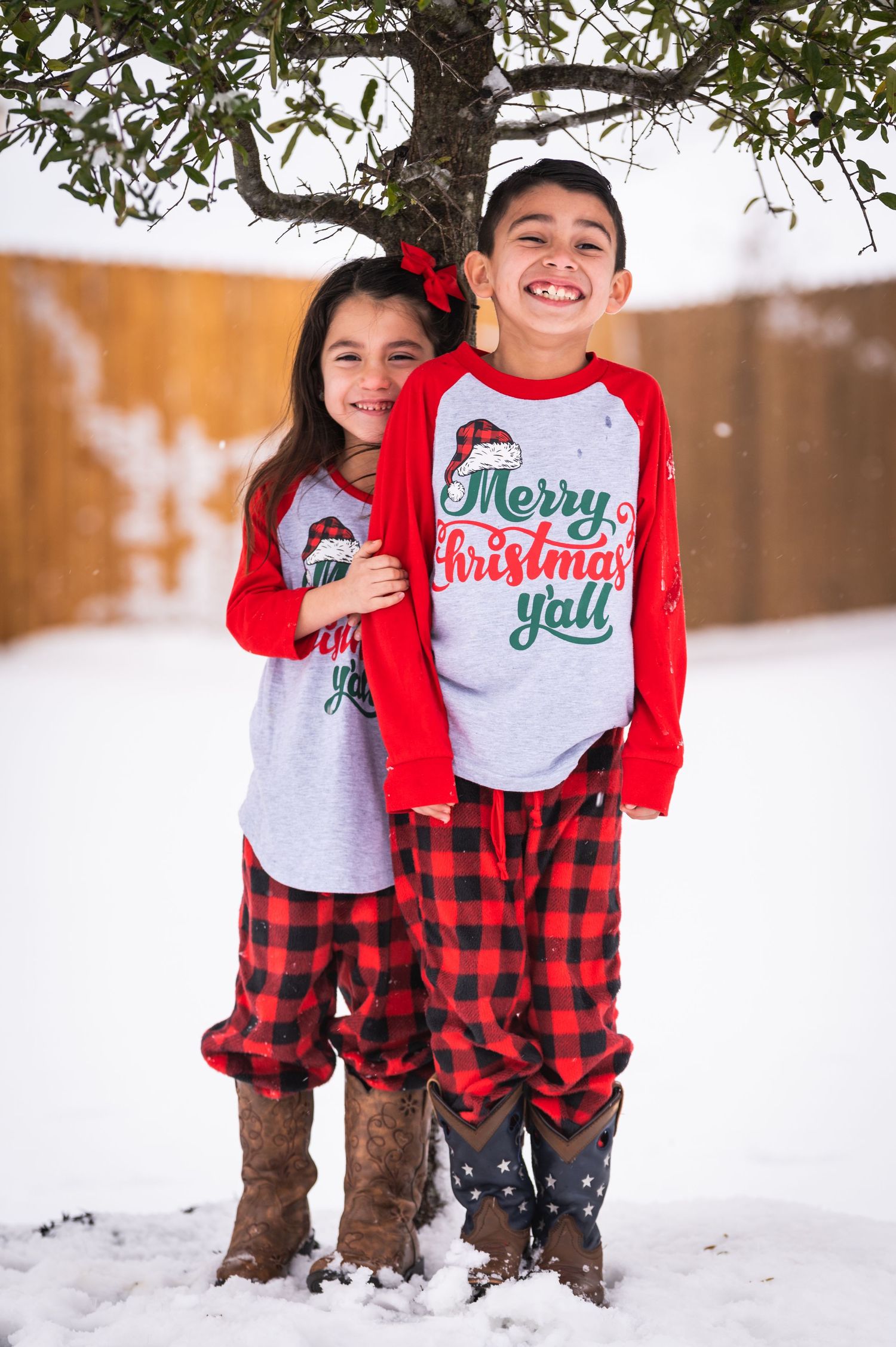 Two children in matching Christmas shirts and plaid pants pose under a tree in the snow.