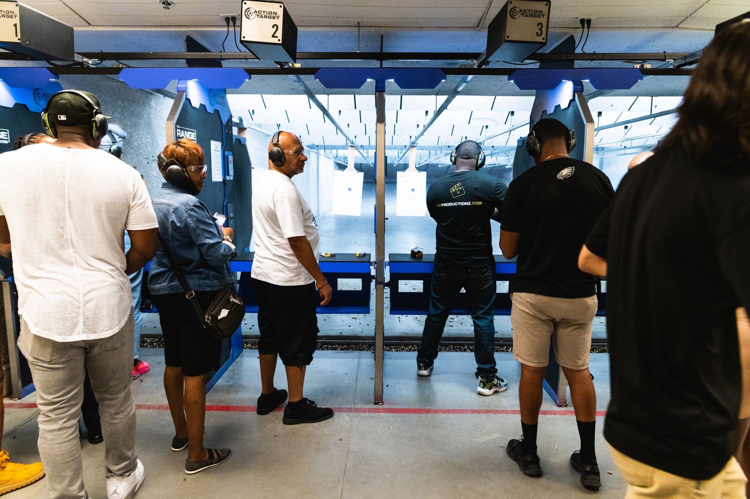 People observing others at an indoor shooting range while wearing protective gear.