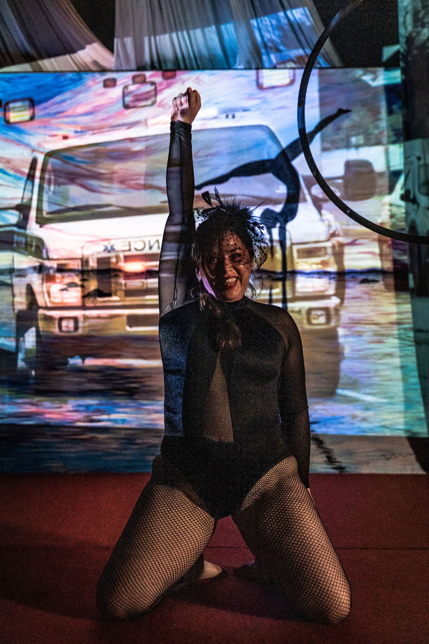 A performer in a black leotard and fishnet stockings kneels on stage against a colorful projected backdrop.