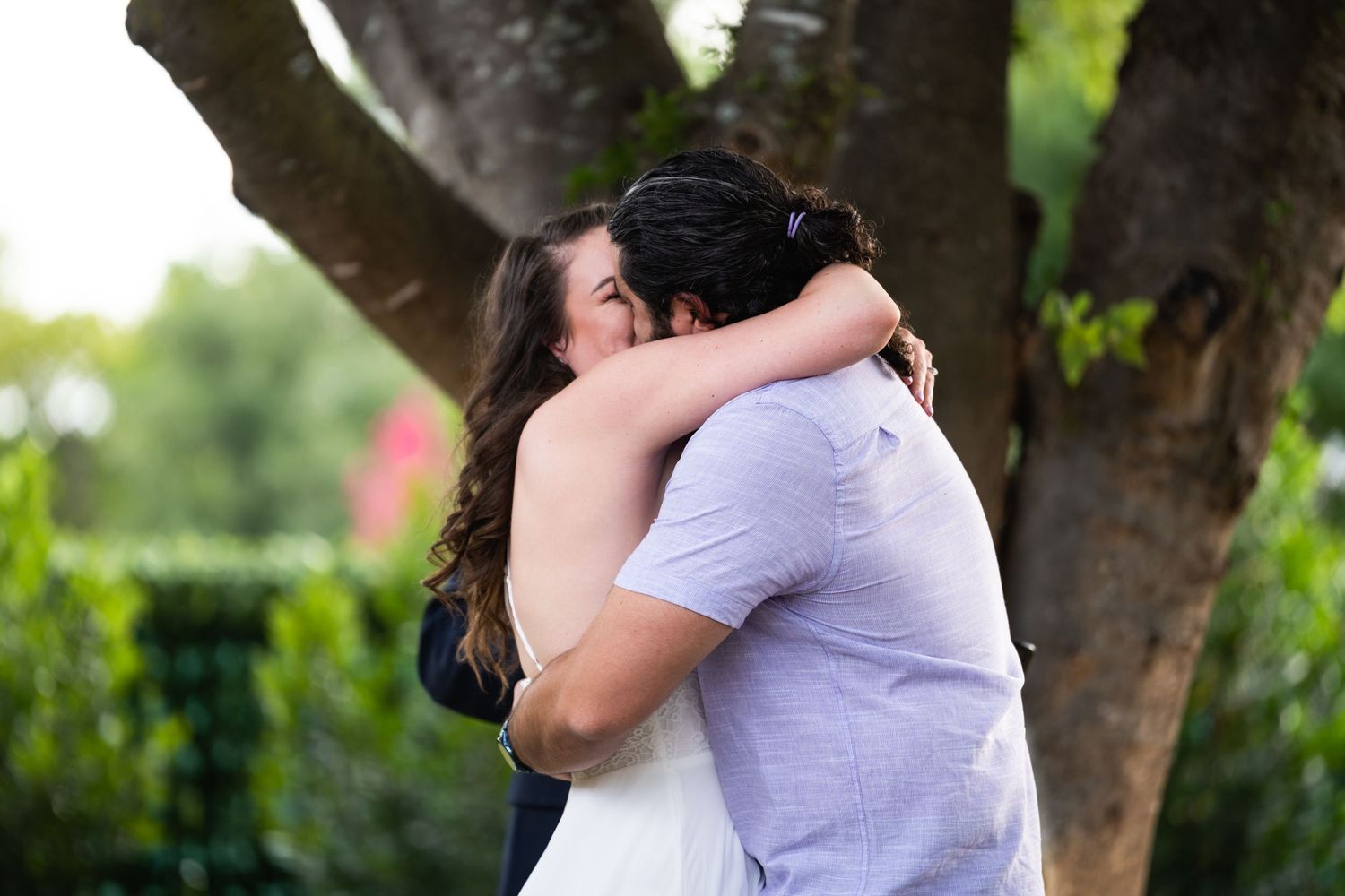 Two people share an intimate embrace outdoors under a tree in a romantic garden setting.