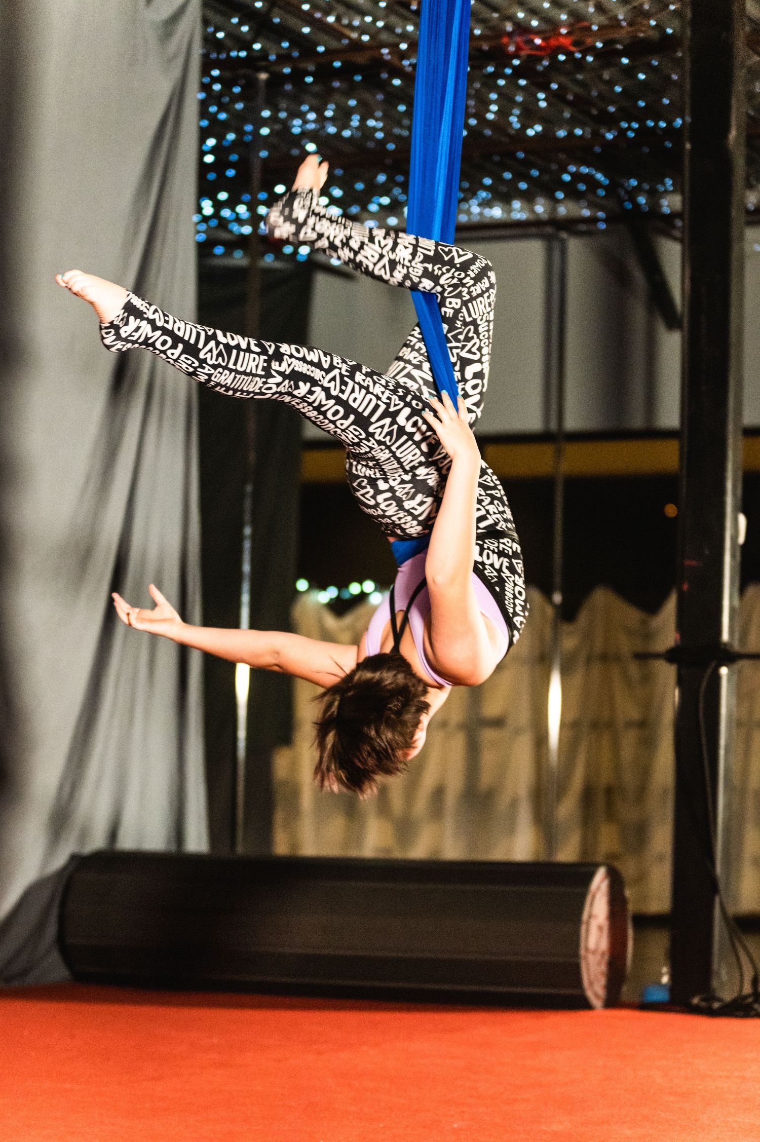 Performer executes inverted split on blue aerial silk against backdrop of twinkling lights.