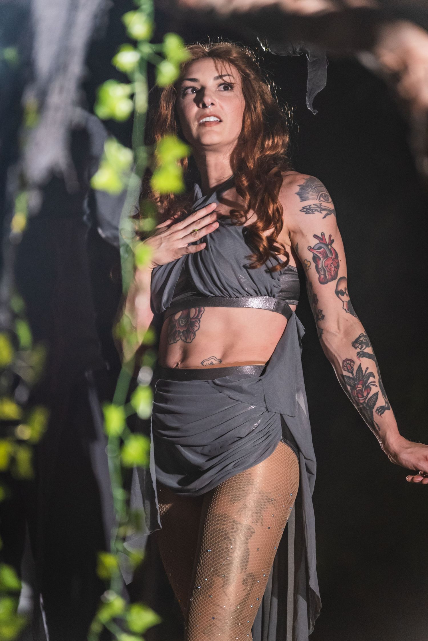Performer in flowing grey costume with tattoos dances dramatically against dark backdrop with leaves.