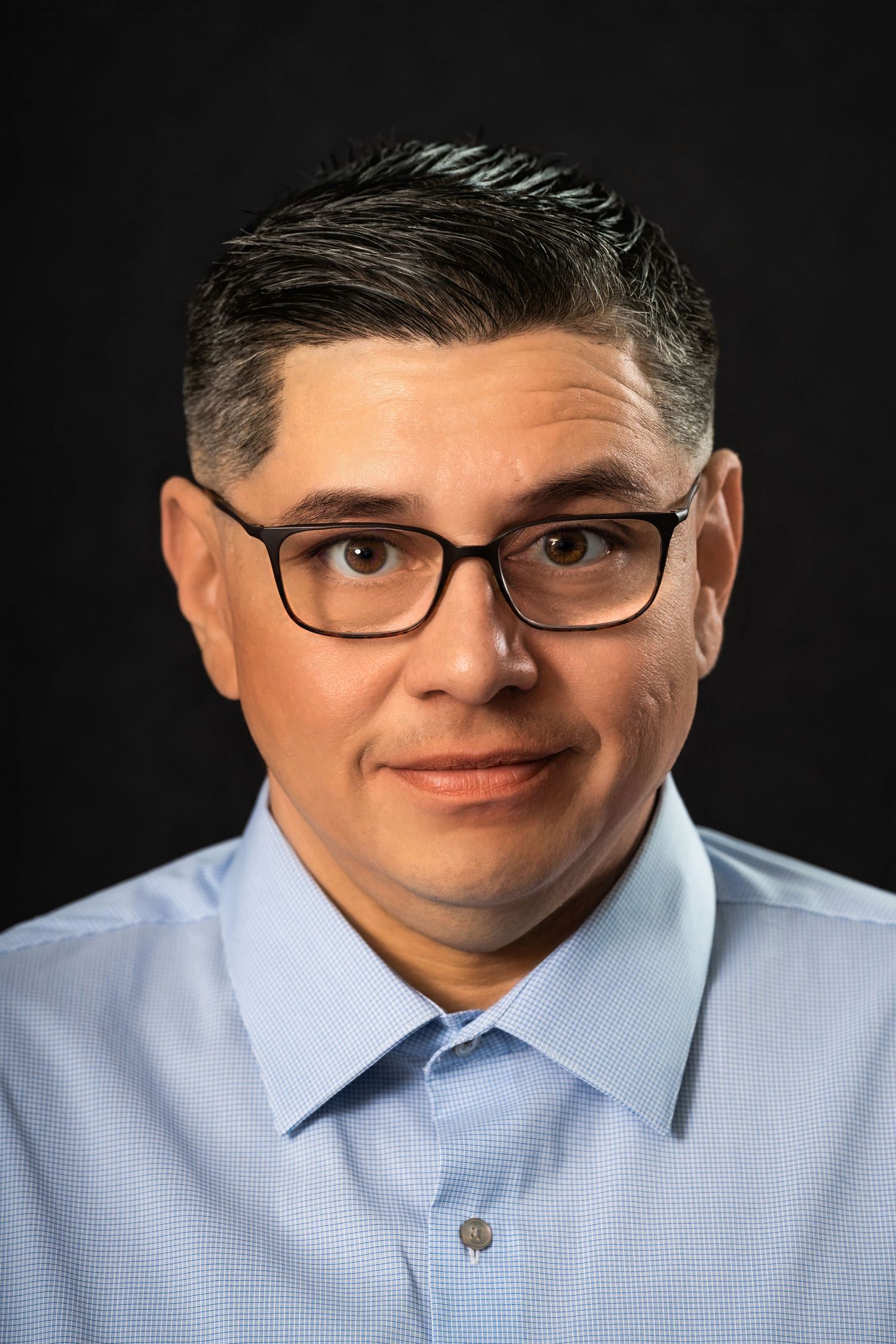 Professional headshot of person wearing glasses and light blue dress shirt against dark background.