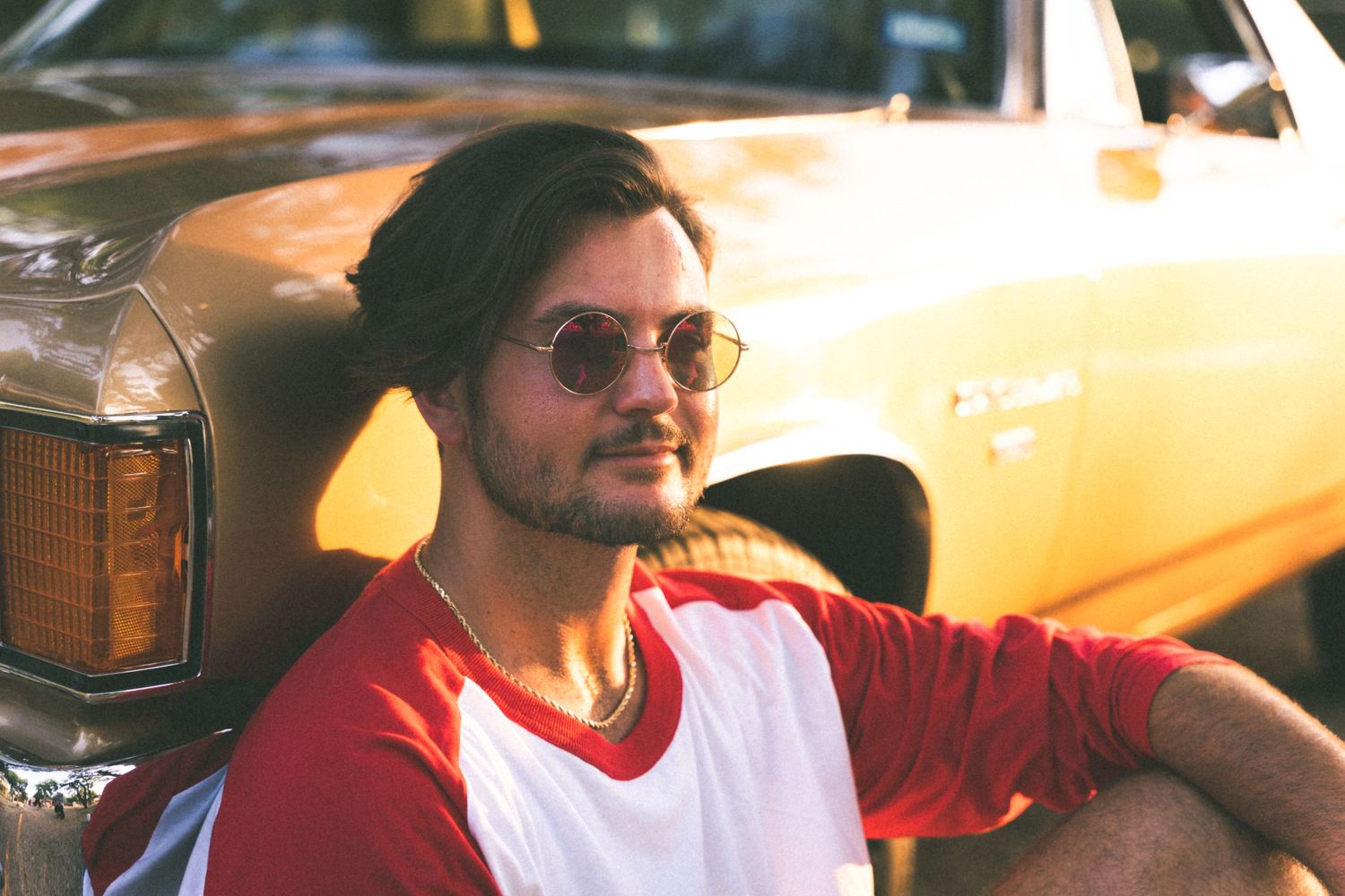 A person in sunglasses and a red and white shirt poses next to a vintage yellow car.