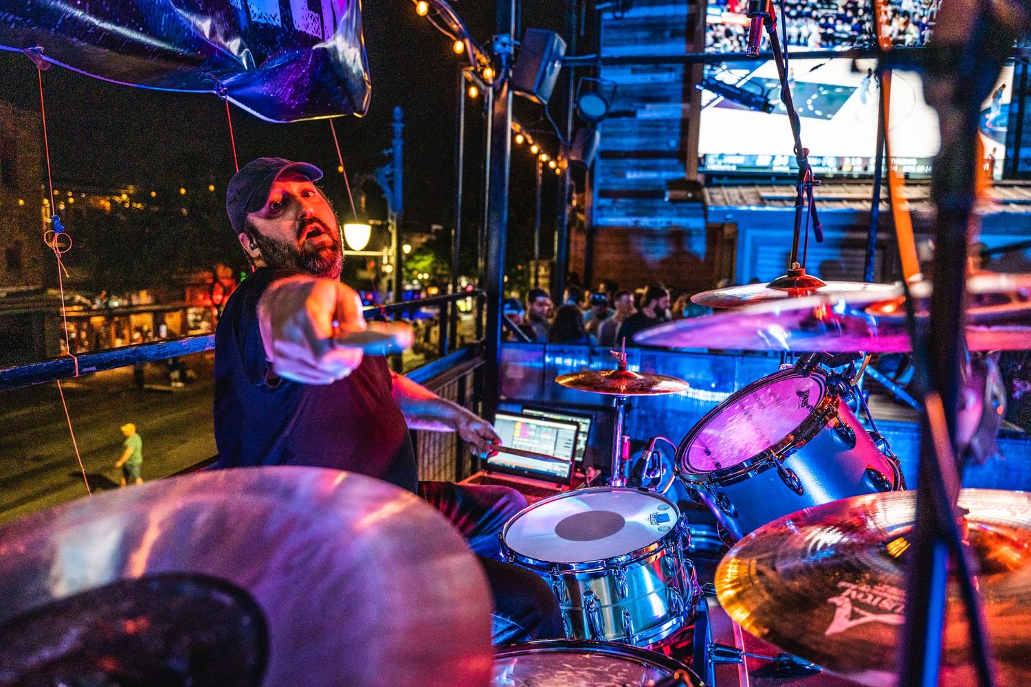 Drummer performing behind drum kit on stage with colorful concert lighting.