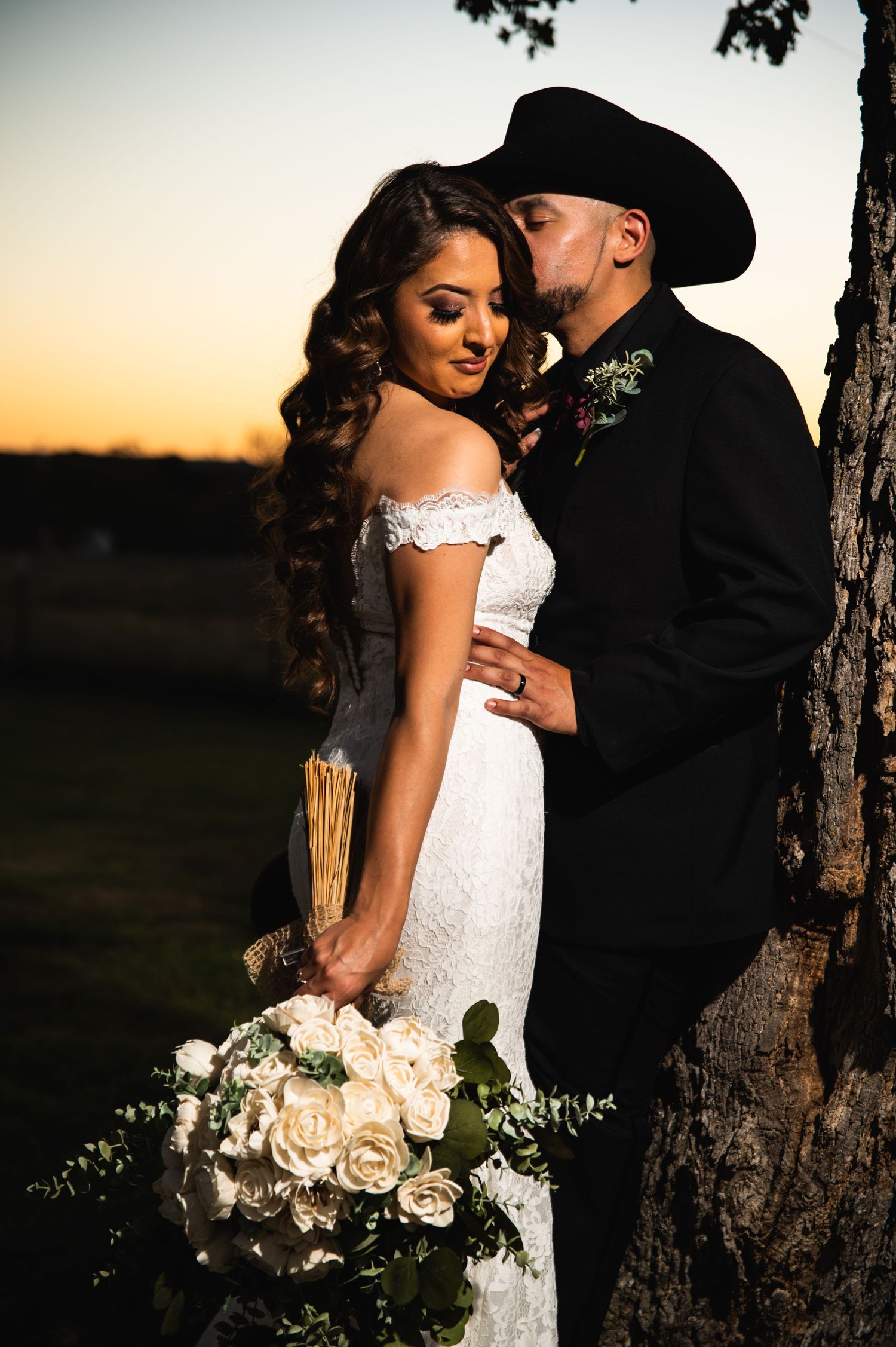 Romantic sunset wedding portrait with cowboy-themed groom and bride holding white rose bouquet by a tree.
