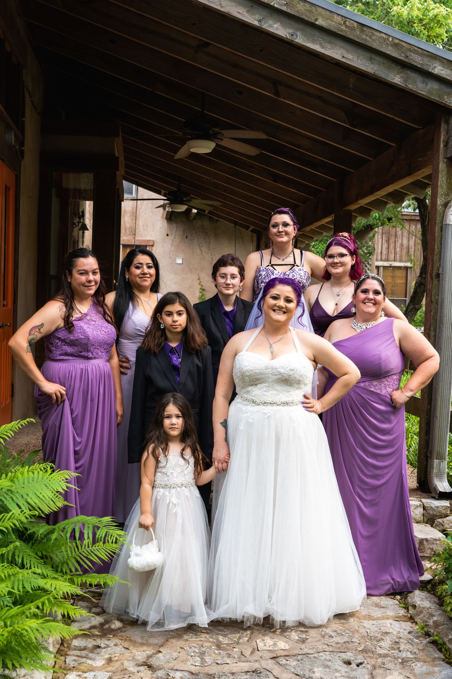A wedding party poses in purple bridesmaid dresses and white gowns under a covered porch.
