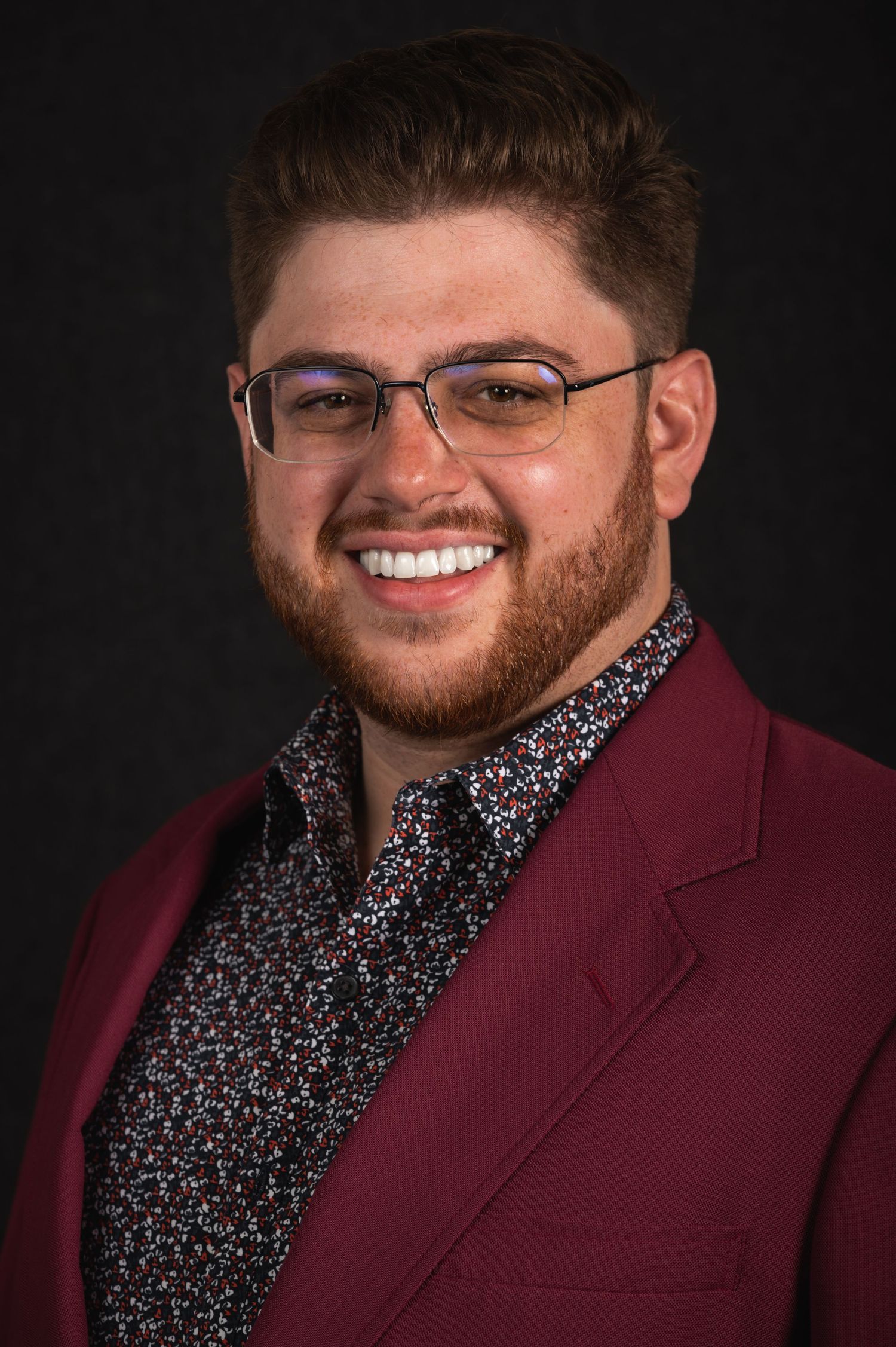 Portrait of a person in glasses and burgundy blazer with patterned shirt smiling at camera.