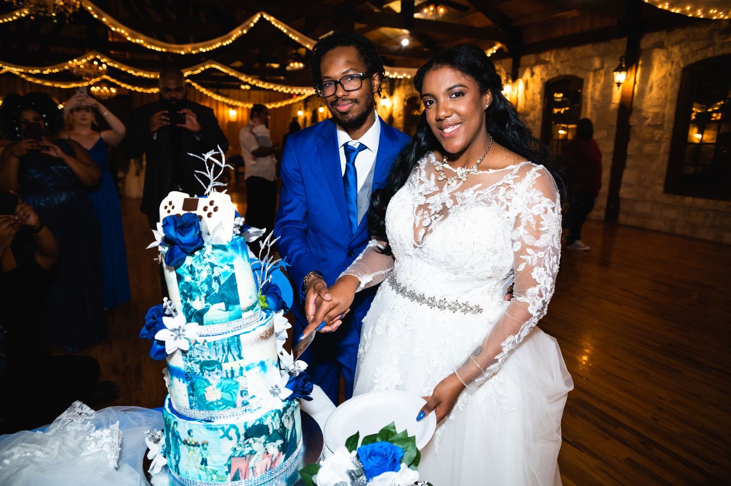 Wedding couple cutting elegant blue and white tiered cake at reception with string lights.
