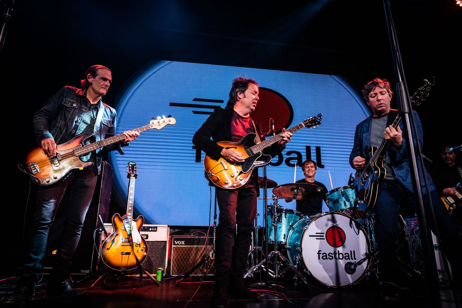 Rock band performs energetically on stage with multiple guitarists and drum kit under blue lighting.
