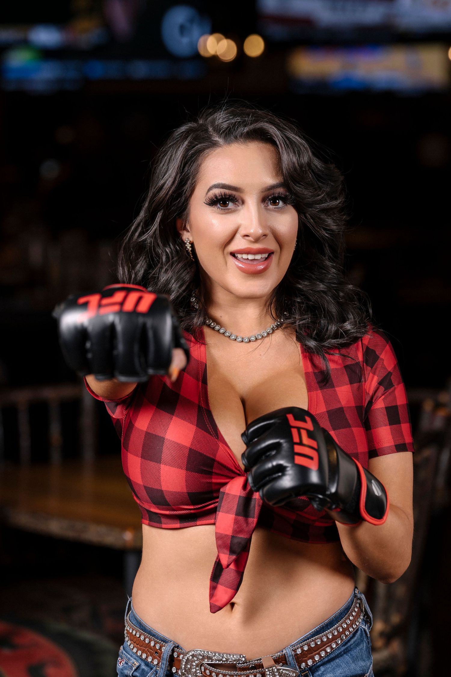Model wearing red plaid crop top shows off UFC gloves in bar setting.