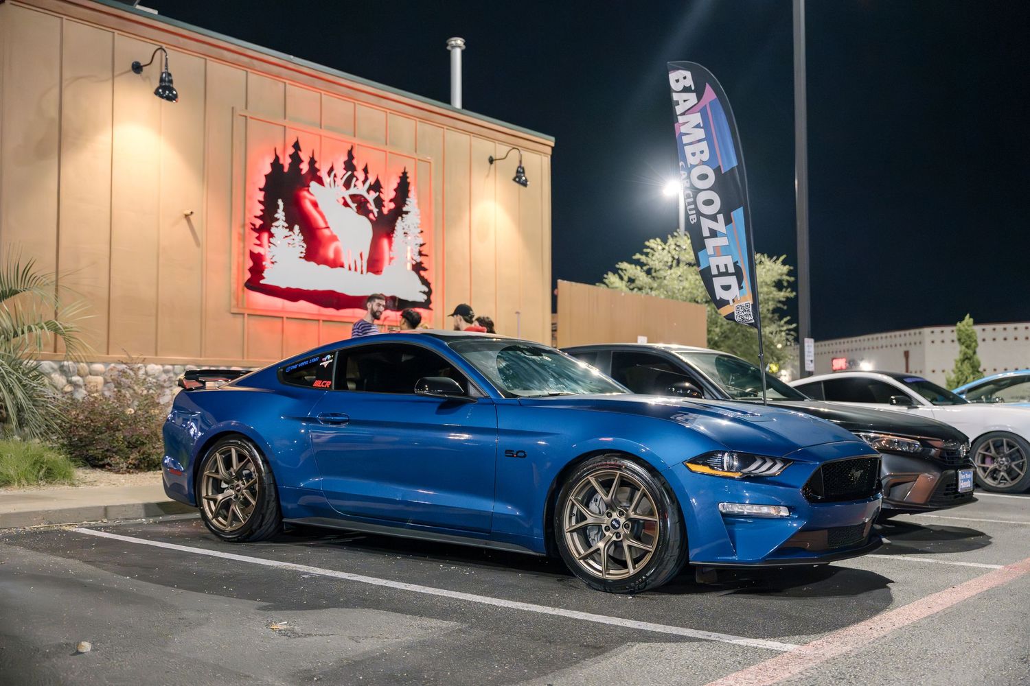 Blue Ford Mustang sports car parked at night with illuminated wall display in background.